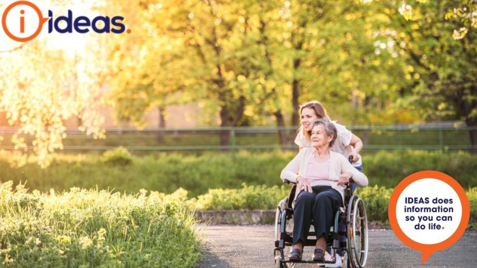Elderly lady in a wheelchair being pushed by her carer. Ideas logo in the image.
